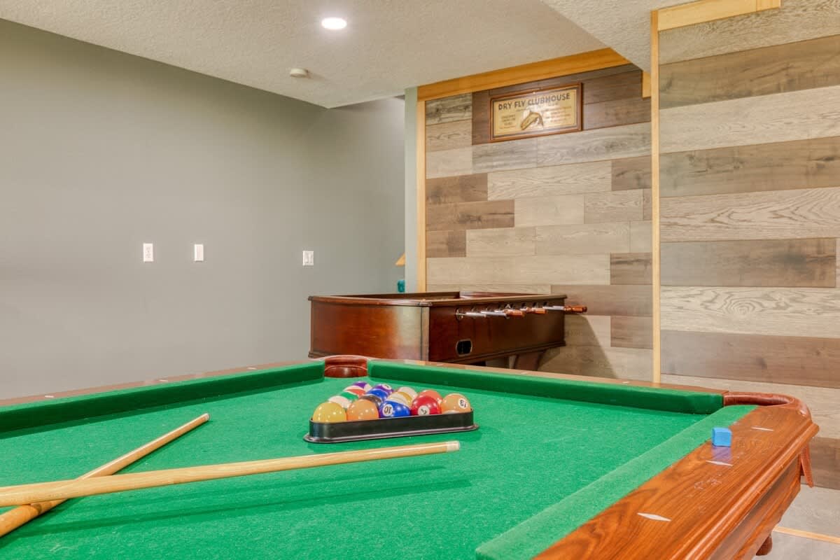 A view of the game room highlighting a green-felted pool table with cues and balls, and a wooden foosball table in the background, set against a wall with decorative wood paneling.