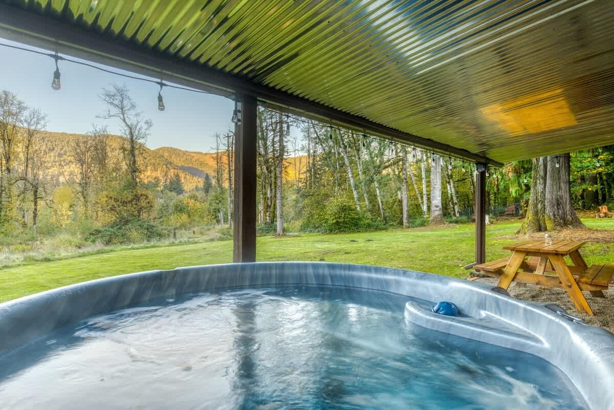 A close-up view of the bubbling hot tub water, offering a serene perspective of a vast green lawn, distant mountains, and a dense forest under a clear sky, with a wooden picnic table visible nearby.