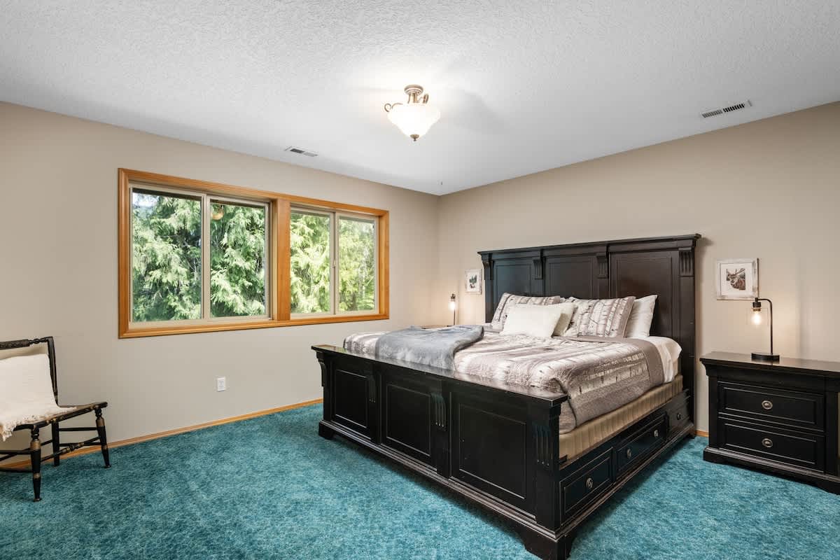 A spacious bedroom featuring a large dark wood king-sized bed with a tall headboard, dressed in patterned bedding. Two large windows with wooden frames offer views of green trees, and the floor is covered in teal carpet.