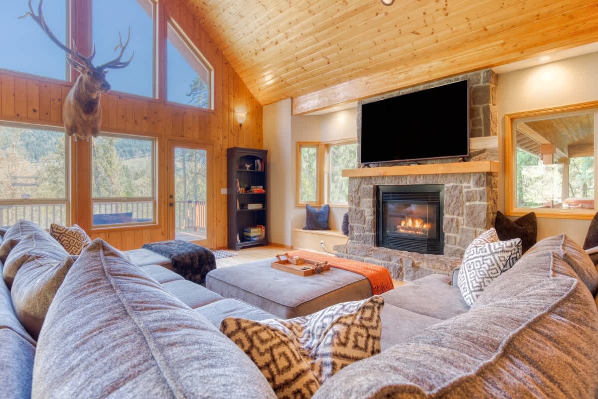A cozy living room with a large grey sectional sofa, a stone fireplace with a mounted TV, and a prominent deer head trophy above the windows, all under a vaulted wooden ceiling.