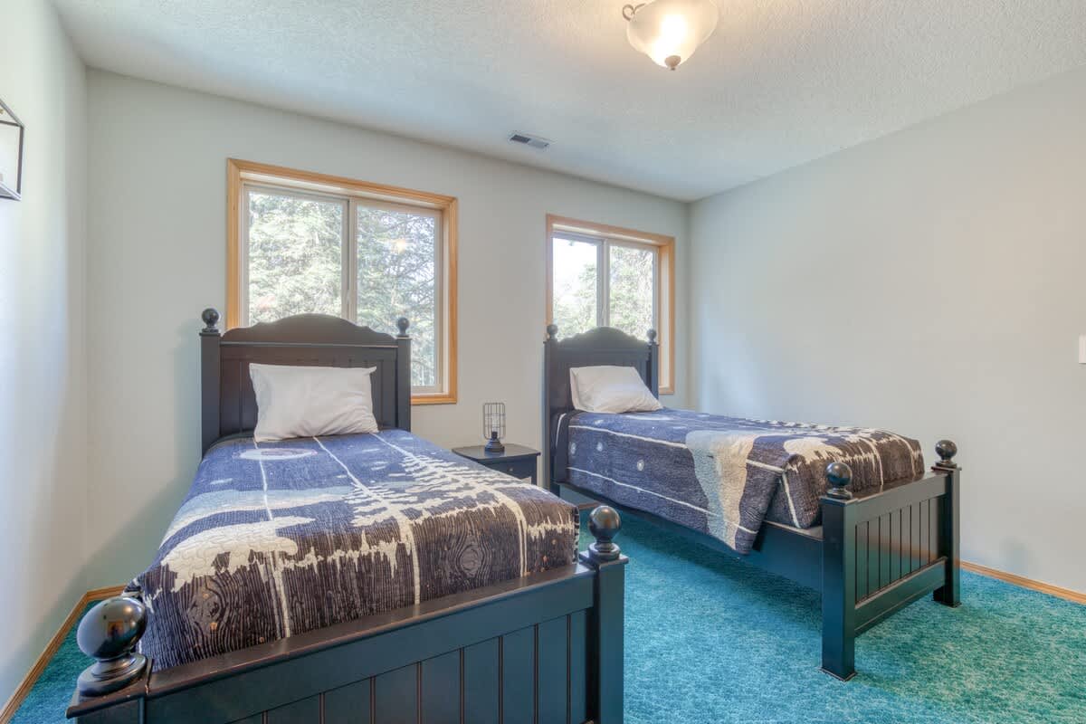 Another view of a bedroom with two dark wood twin beds, each with different blue and white patterned bedding. The room has light grey walls, teal carpet, and two wooden-framed windows overlooking trees.