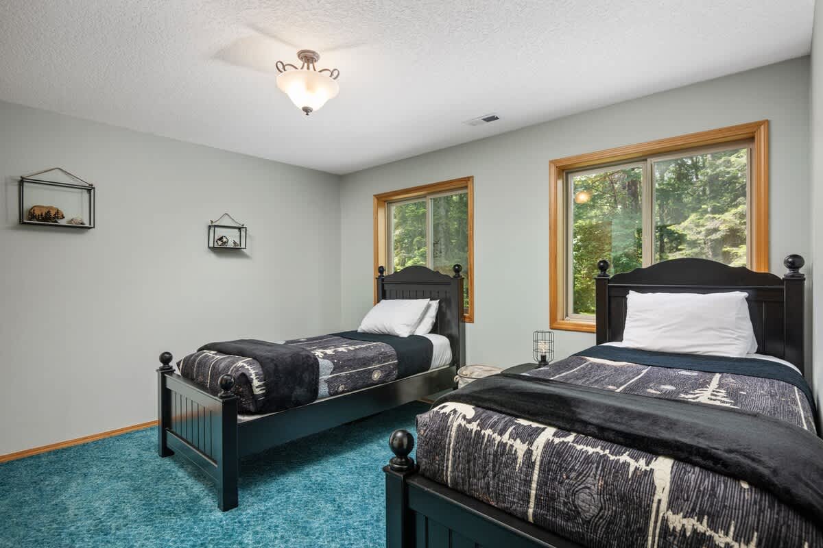 A bright bedroom featuring two dark wood twin beds with black and white patterned bedding, set against light grey walls. Two large wooden-framed windows offer views of green trees, and the floor is teal carpet.