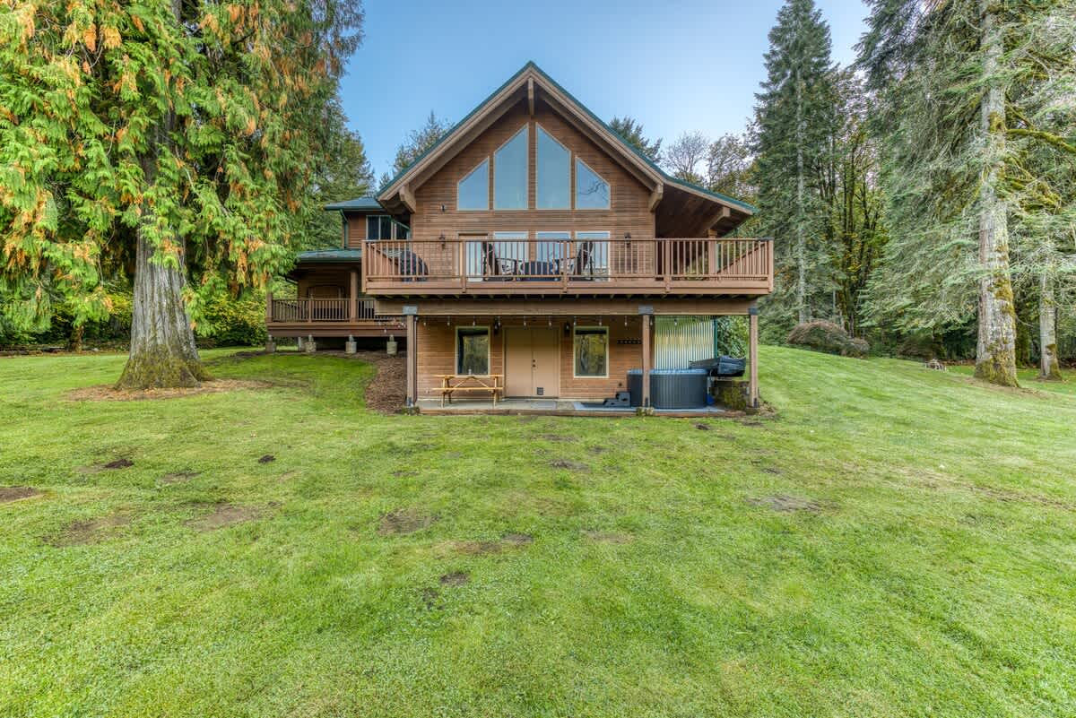 The back exterior of a rustic wooden lodge with a prominent A-frame section, large windows, and an expansive upper deck, with a hot tub and picnic table on the lower level, set against a backdrop of tall trees.