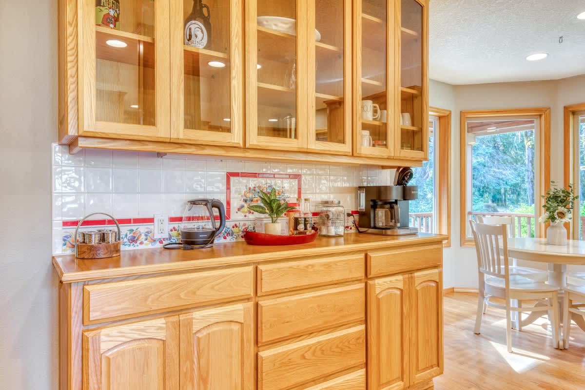 Dedicated coffee bar area with light wood base and glass-front upper cabinets, white tiled countertops, and a decorative tiled backsplash. A coffee maker, kettle, and various serving items are neatly arranged on the counter.