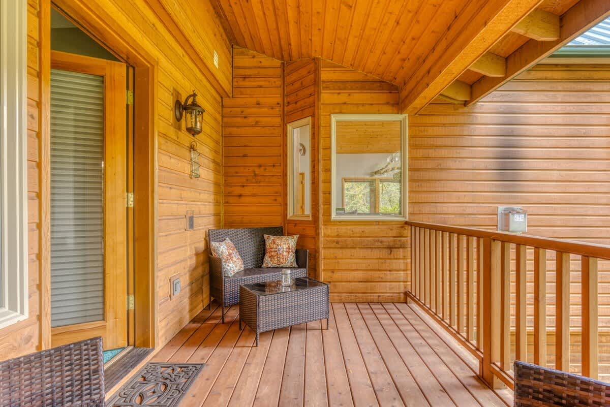 A section of the covered wooden porch featuring a cozy wicker seating area with two chairs and a coffee table. The porch is characterized by its natural wood paneling and railing, offering a glimpse of the forest.