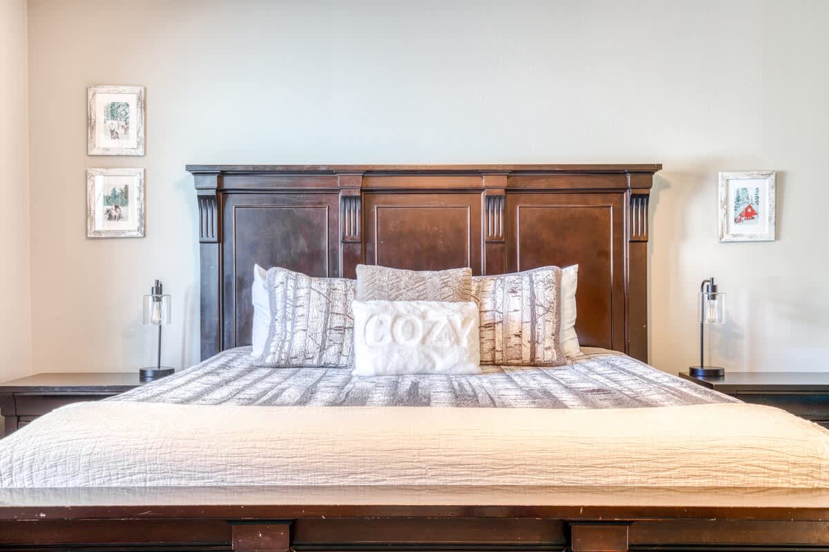 Close-up of a dark wood king-sized bed with a tall, ornate headboard, featuring white and grey patterned bedding and a "COZY" throw pillow. Two small lamps sit on dark nightstands flanking the bed.