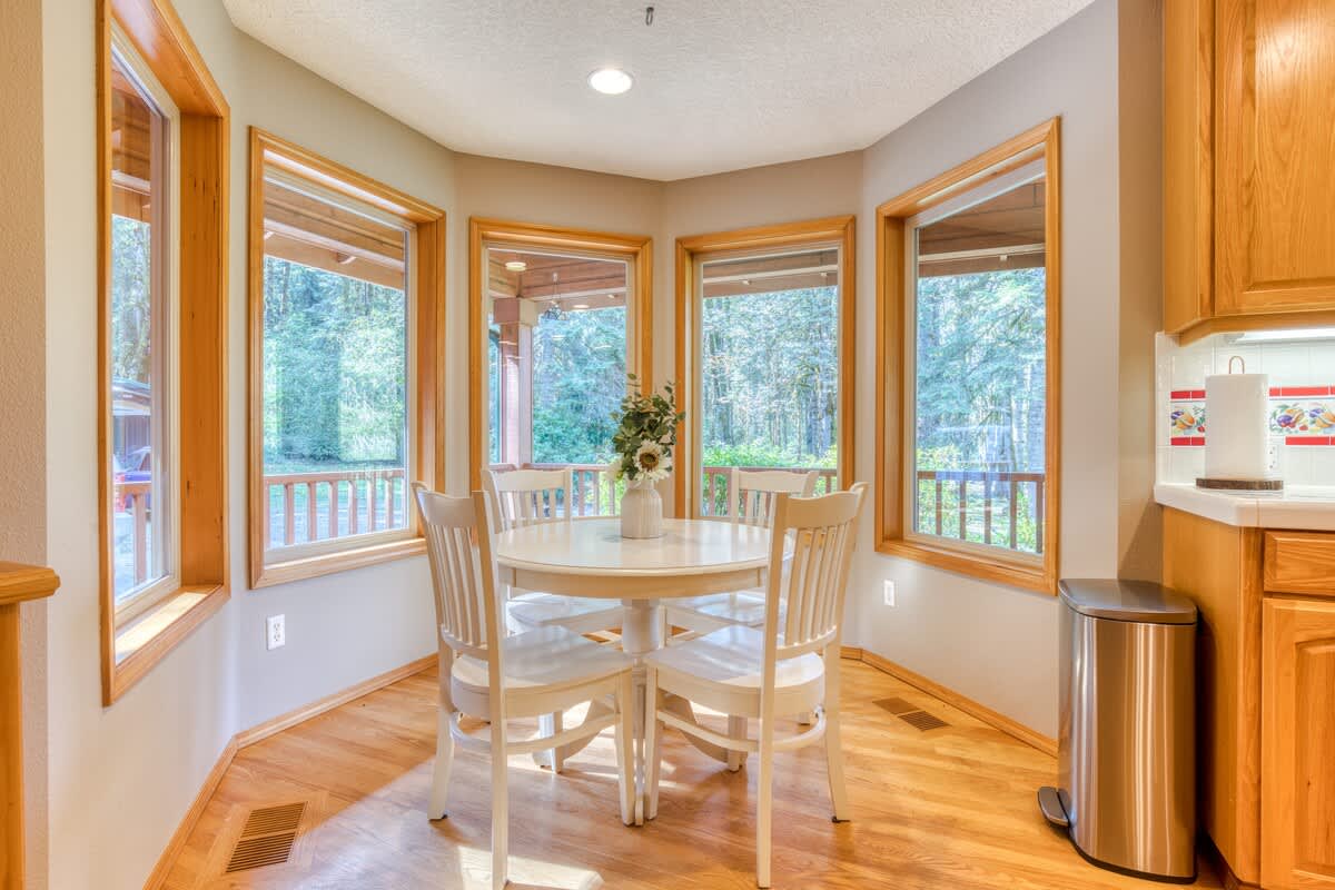 Bright breakfast nook with a round white dining table and four matching chairs, surrounded by large bay windows offering views of the outdoor greenery. Light wood cabinetry is visible to the right.