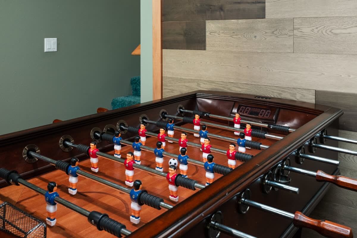 Close-up of a dark wood foosball table with red and blue players, featuring a digital scoreboard and a soccer ball in play, set against a wall with wood paneling and a green accent.