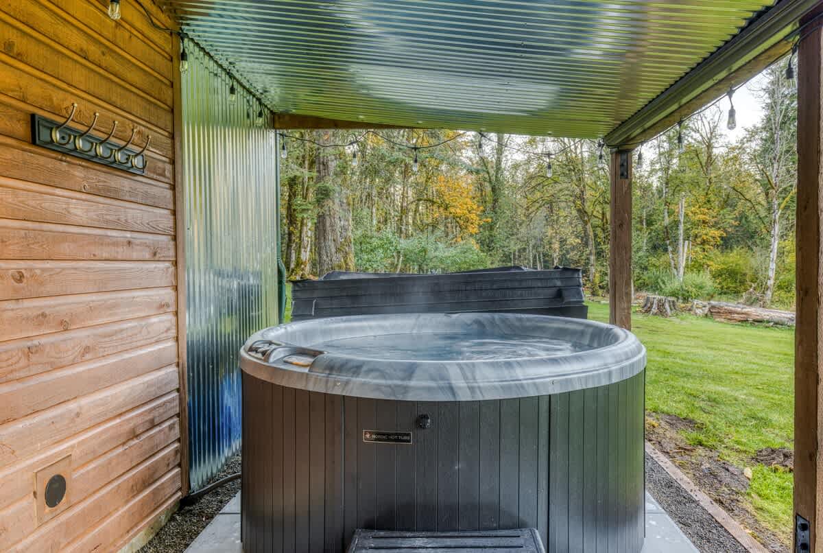 A large, dark grey hot tub is situated under a corrugated metal roof, next to a wooden-sided house, with a lush green forest visible in the background.