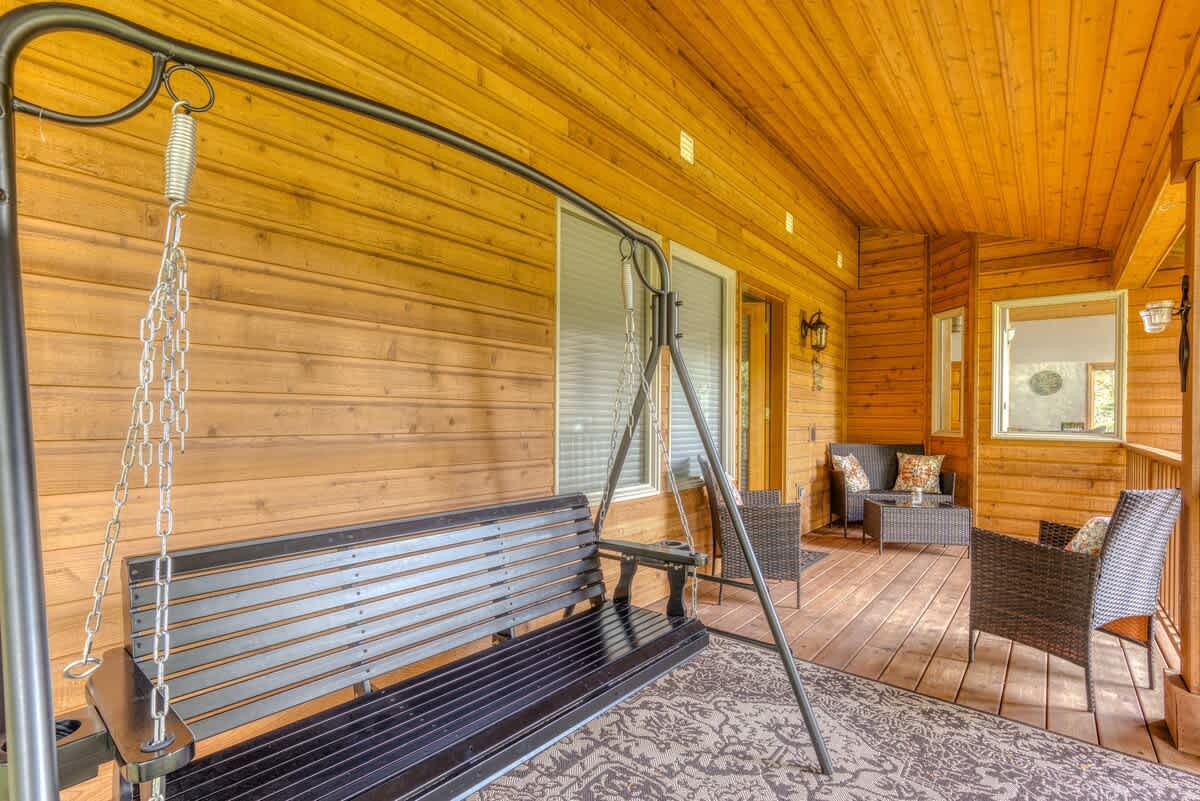 A wide shot of the covered wooden porch, featuring a black metal swing bench, two wicker chairs, and a small table. The porch boasts a warm wood-paneled ceiling and walls, with a view of the surrounding forest.