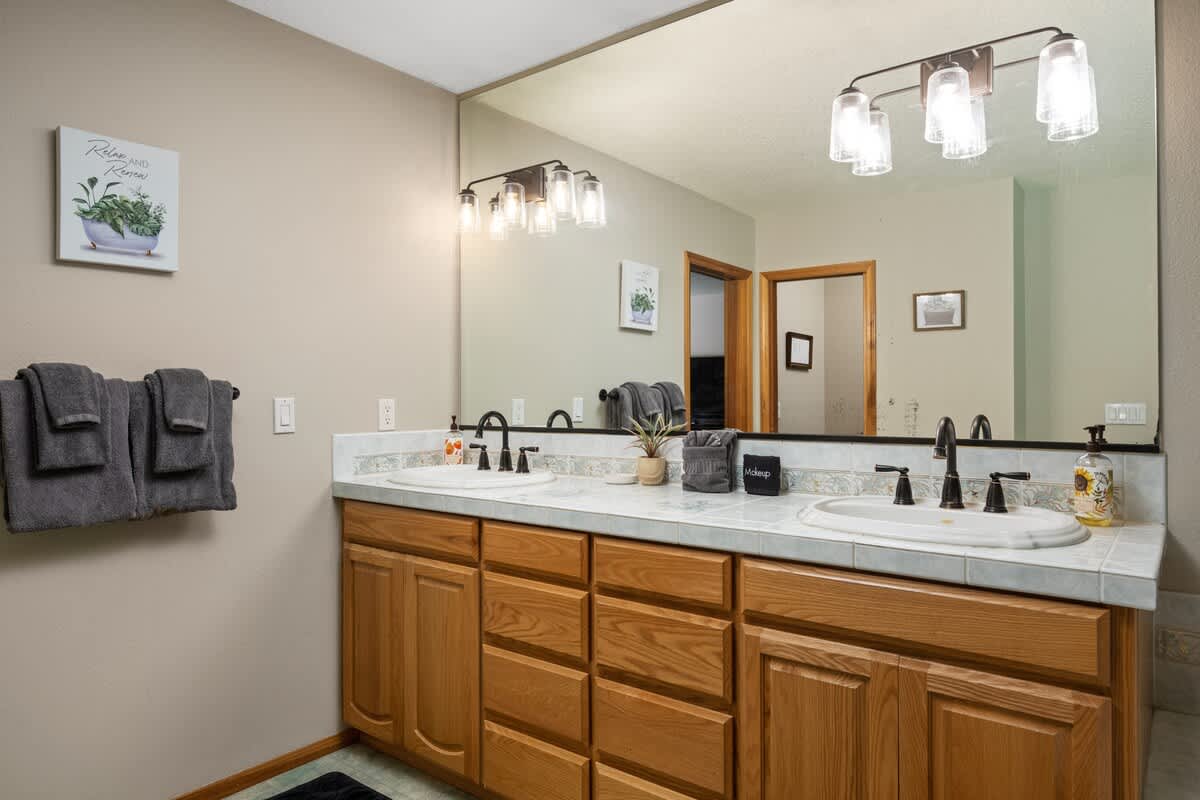 A spacious bathroom with a long double vanity featuring light wood cabinets, a tiled countertop, and two white oval sinks with black faucets. A large mirror spans the wall above, reflecting the room and a doorway.