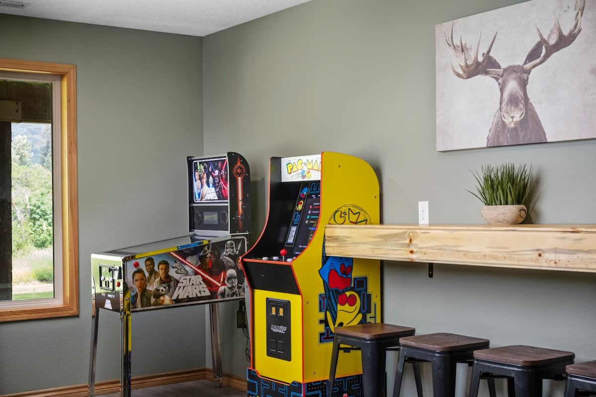 A close-up of the game room's arcade corner, showcasing a Star Wars pinball machine and a classic yellow Pac-Man arcade cabinet, next to a wooden bar with metal stools.