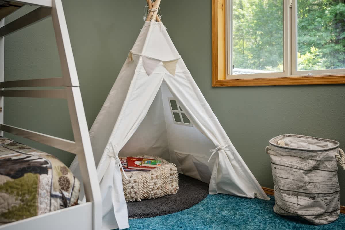A close-up of a white canvas teepee with small window cutouts, positioned on a dark circular rug next to a white wooden bunk bed, with a woven basket nearby.