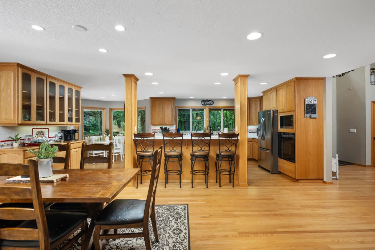 Open-concept kitchen and dining area featuring light wood cabinetry, hardwood floors, and a large island with bar seating. A wooden dining table with chairs is visible in the foreground, and large windows offer views of the outdoors.