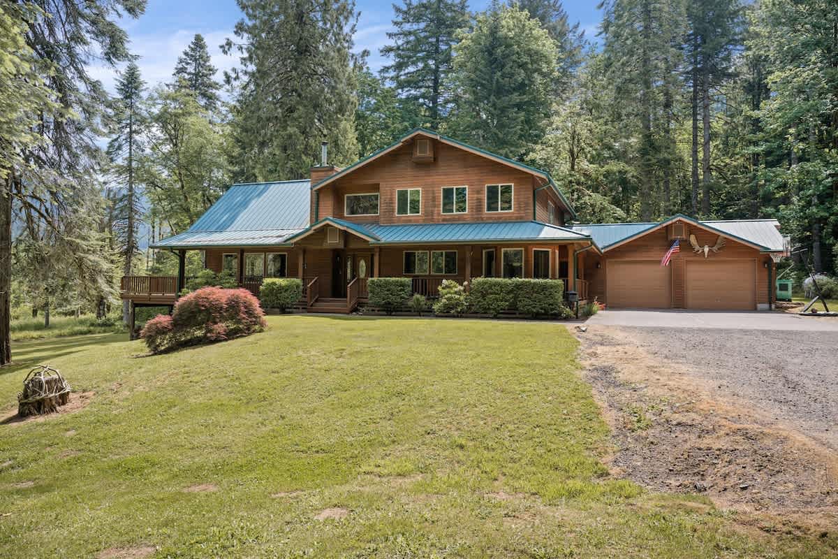The front of a large, rustic wooden house with a green metal roof is seen from a ground-level perspective, featuring a gravel driveway leading to the garage and surrounded by a well-maintained lawn and tall trees.