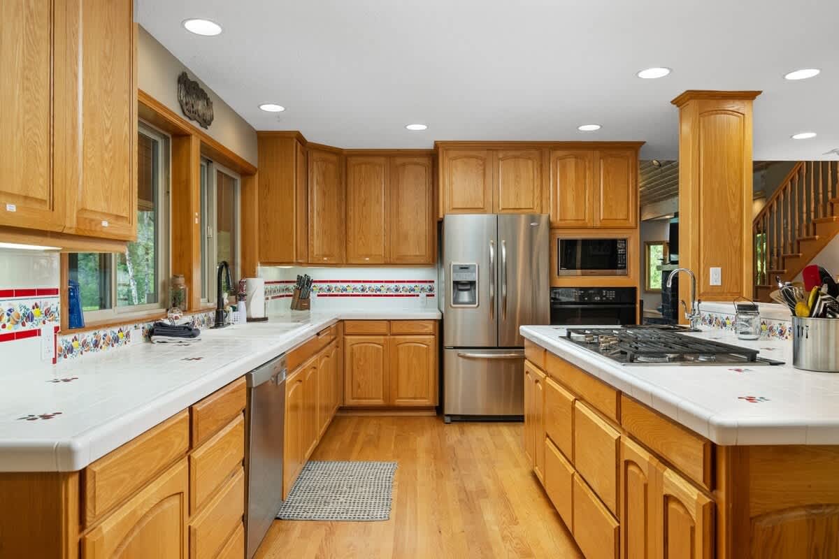 Well-equipped kitchen featuring light wood cabinetry, white tiled countertops, and stainless steel appliances including a refrigerator, dishwasher, and built-in oven. A gas cooktop is integrated into the island, and large windows overlook a wooded area.