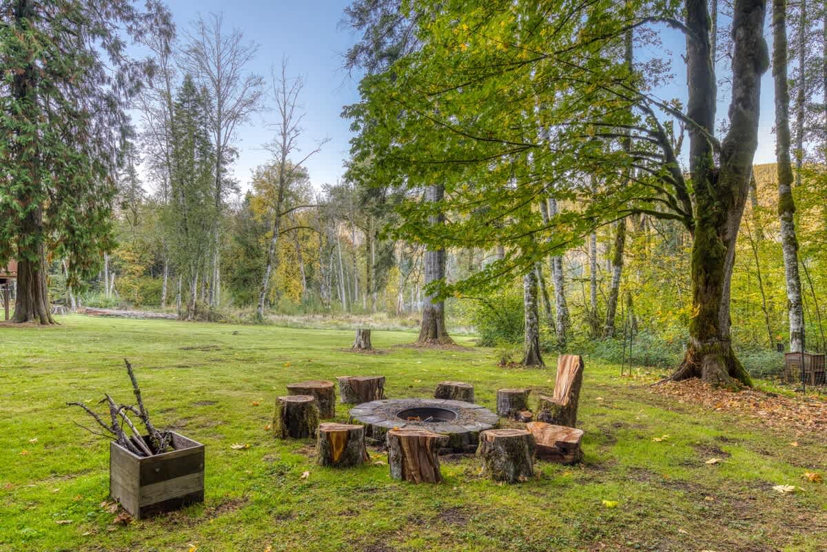 An inviting outdoor fire pit area featuring a circular arrangement of tree stump seats around a stone fire pit, with a wooden chair and log holder nearby, set in a grassy clearing surrounded by trees.