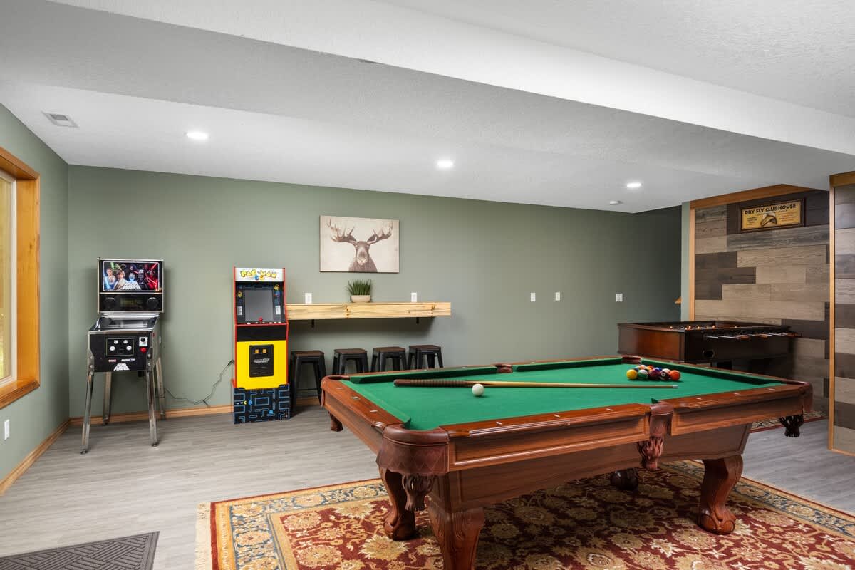 A vibrant game room featuring a pool table, a Pac-Man arcade machine, a Star Wars pinball machine, and a long wooden bar with stools, all against olive green walls.