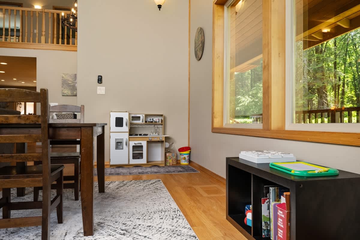 A dedicated children's play area within the living space, featuring a white wooden play kitchen and a black low bookshelf with games and books. Large windows offer natural light, and light hardwood floors are visible.