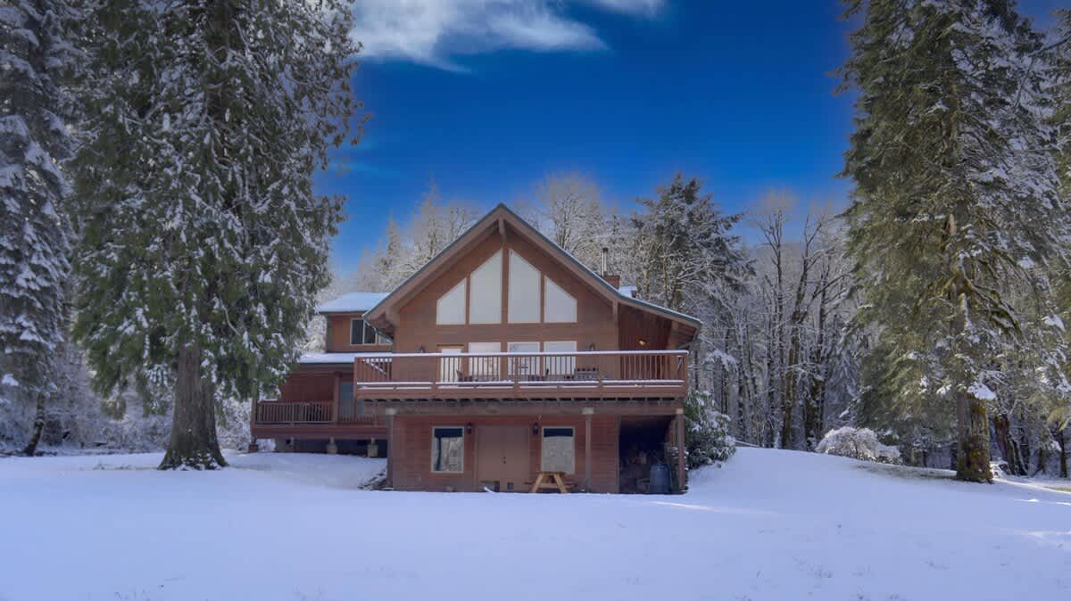The back exterior of the wooden lodge in winter, featuring its A-frame design, large windows, and upper deck, all covered in a blanket of snow, with snow-laden evergreen trees in the background under a clear blue sky.