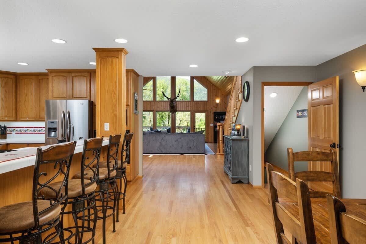 An open-concept kitchen and dining area with light wood cabinetry, stainless steel appliances, and a kitchen island with bar stools. A wooden dining table is in the foreground, and the living room with large windows is visible beyond.