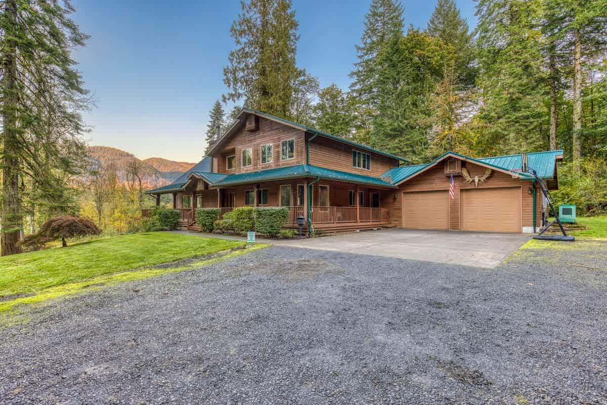 Large, rustic wooden lodge with a teal metal roof, a wide front porch, and an attached two-car garage, set amidst tall evergreen trees with mountains visible in the distance.