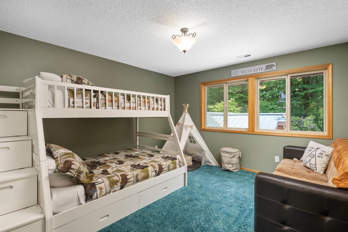 A children's bedroom featuring white wooden bunk beds with a lower full bed and an upper twin, a white teepee, and a dark teal carpet, all set against olive green walls. A small black sofa is visible on the right.