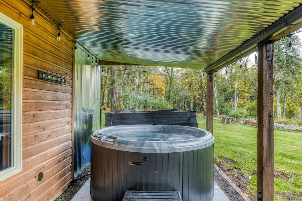 A round, dark-colored hot tub situated under a corrugated metal roof covering, with a view of a grassy yard and dense forest in the background, next to the wooden siding of the house.