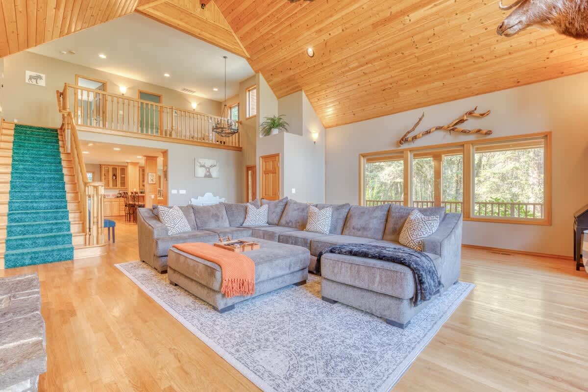 A wide view of a rustic living room featuring a large gray sectional sofa, an ottoman with a blue throw, light hardwood floors, and a vaulted wooden ceiling. A teal carpeted staircase leads to a mezzanine level with a wooden railing.