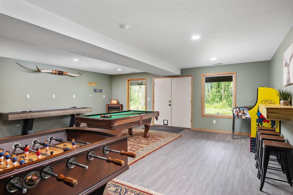 A spacious game room with light green walls features a foosball table in the foreground, a pool table, a shuffleboard table, and a classic arcade machine, all on a mix of wood-look flooring and area rugs.