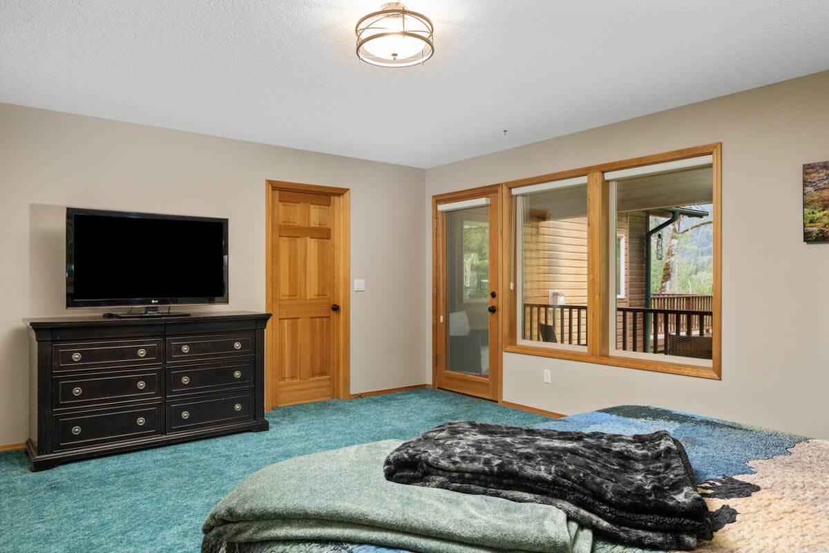 Another view of the bedroom showcasing a large flat-screen TV on a dark wooden dresser, a natural wood door, and a glass door leading to an outdoor area. The room maintains its blue carpet and light beige walls.