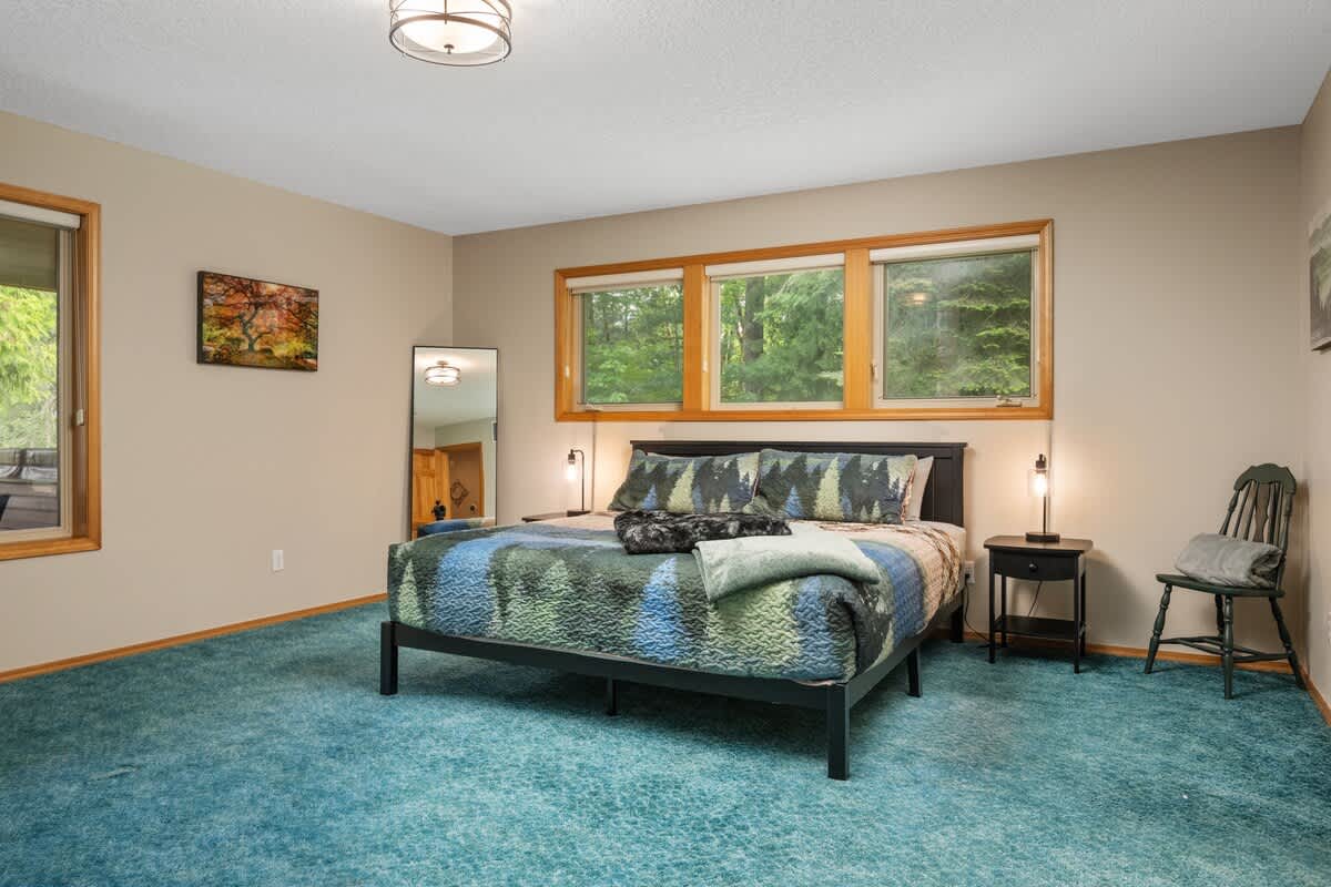 A spacious bedroom with a black metal bed frame, a blue and green patterned comforter, and large windows offering a lush forest view. The room features light beige walls and soft blue carpet.