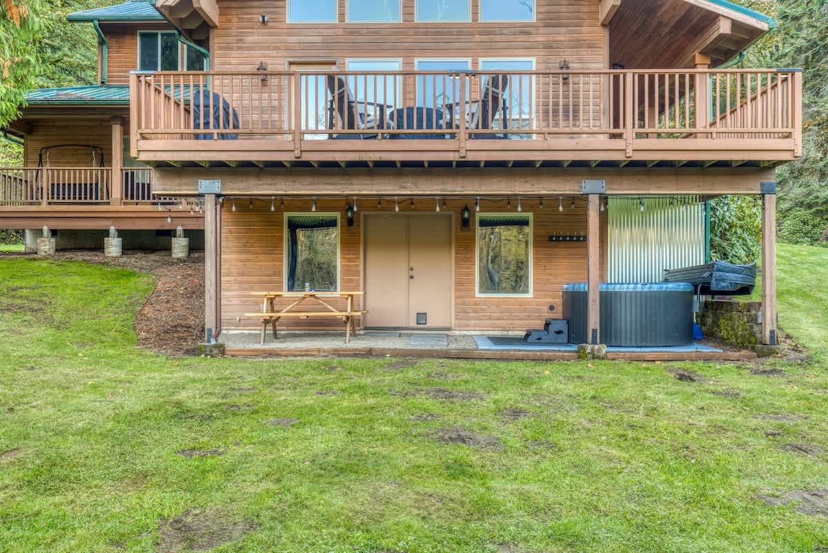 The back exterior of the wooden house, showing the elevated deck above and a ground-level patio area with a hot tub and a wooden picnic table, surrounded by a green lawn and trees.