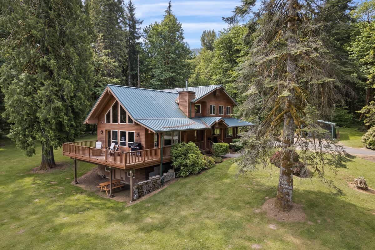An aerial view showcases a large, rustic wooden house with a green metal roof, surrounded by dense green trees and a sprawling lawn, featuring a spacious wooden deck with outdoor seating.