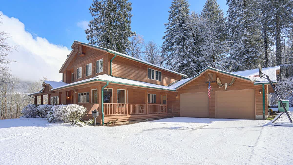 The front exterior of the wooden lodge in winter, with a snow-covered driveway and grounds, showcasing its green metal roof and attached garage, surrounded by snow-dusted evergreen trees under a bright blue sky.
