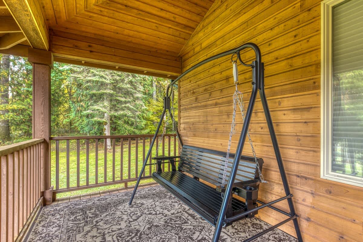 A close-up view of the black metal swing bench on the covered wooden porch, placed on a decorative outdoor rug. The background reveals a lush, green forest.