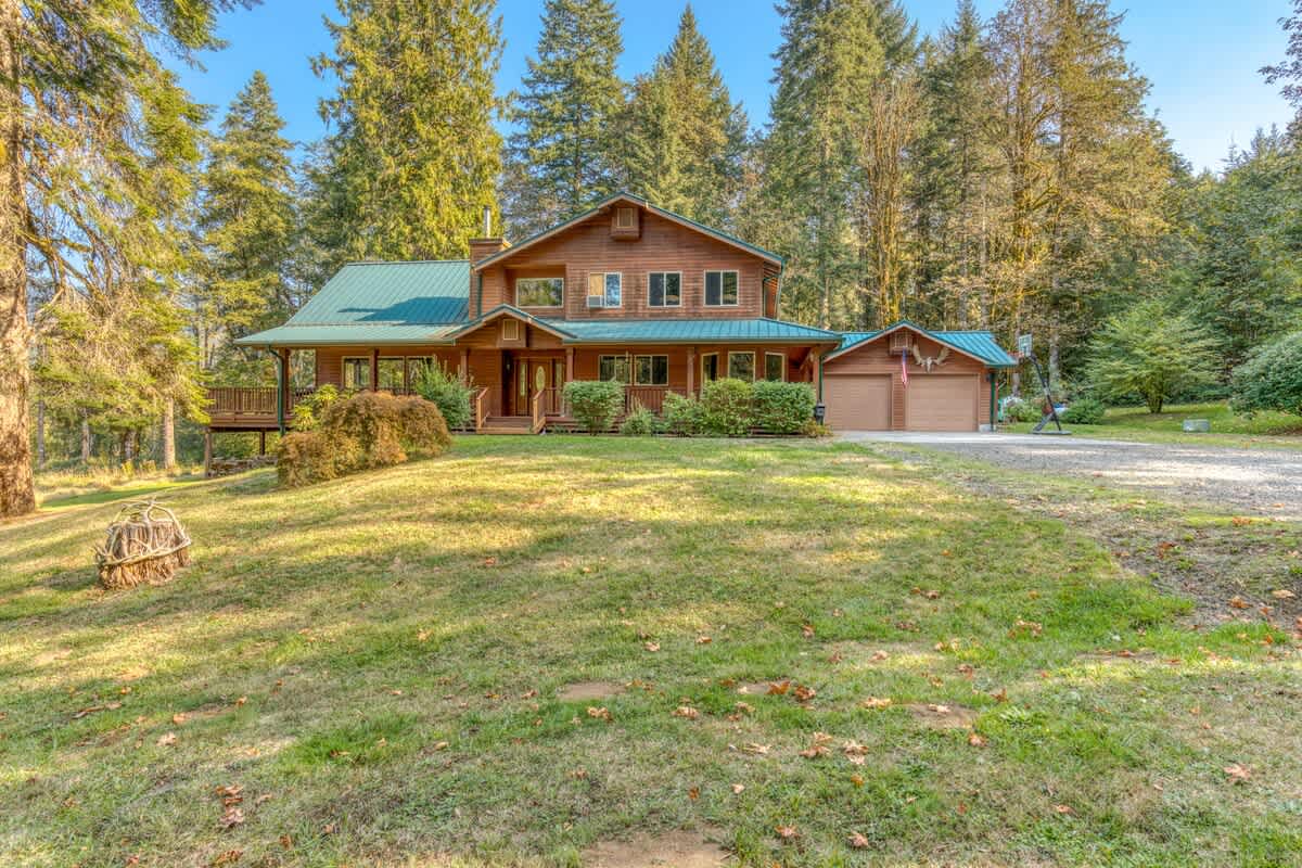 The front exterior of a spacious wooden lodge with a green metal roof and an attached garage, situated on a large grassy lawn with a gravel driveway, surrounded by tall evergreen trees.
