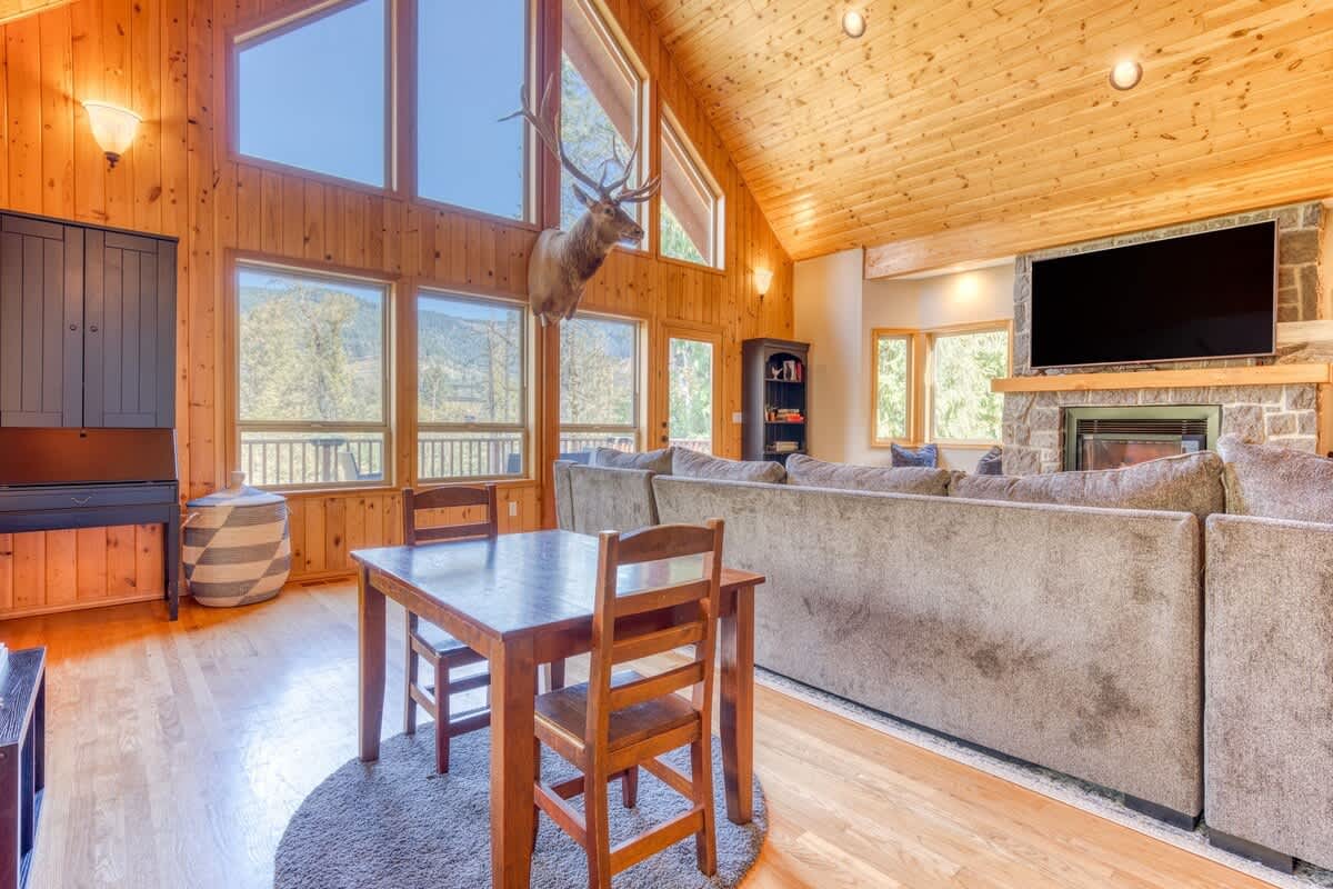 A rustic living area featuring a small wooden dining table with two chairs on a round rug, adjacent to a large gray sectional sofa. The room has light hardwood floors, a vaulted wooden ceiling, and large windows.