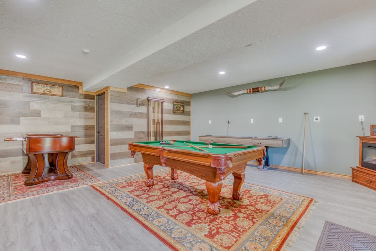 A large game room with a wooden pool table as the centerpiece, surrounded by light gray flooring and accent walls, with a shuffleboard table and a foosball table visible in the background.