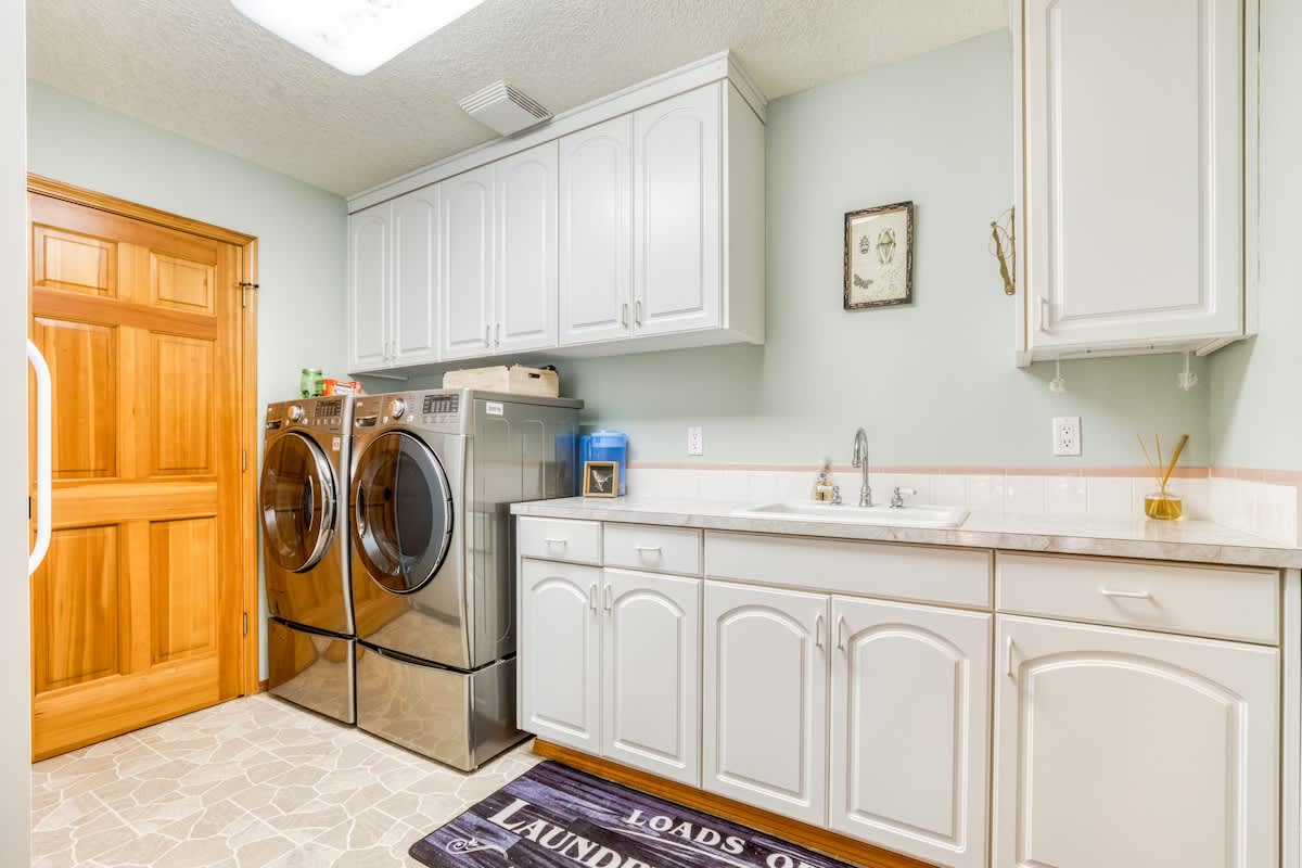 A bright laundry room with light green walls, white cabinetry, a utility sink, and stainless steel front-loading washer and dryer units, featuring a light-colored tiled floor.