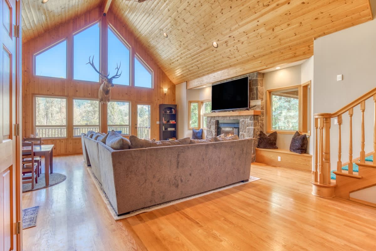 A view from behind a large gray sectional sofa in a living room, showing a stone fireplace with a TV above, and a wooden staircase with teal carpet. The room has light hardwood floors and a vaulted wooden ceiling with large windows.