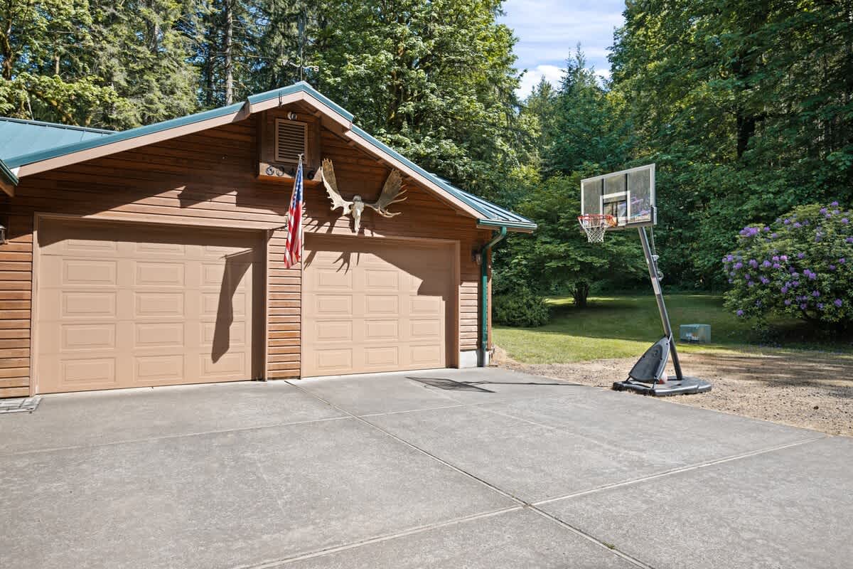 A two-car garage with light brown doors and wooden siding features an American flag and decorative antlers above the doors, with a portable basketball hoop set up on the concrete driveway.