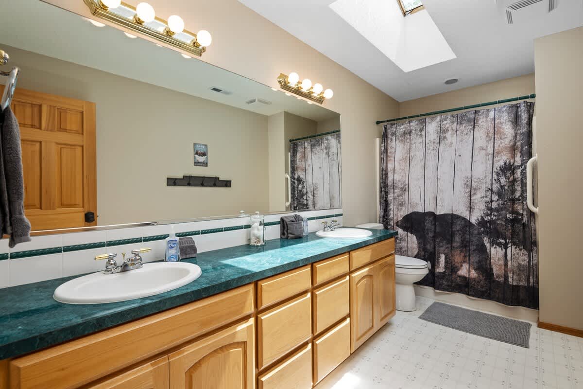 A spacious bathroom featuring a long wooden vanity with double sinks and a green countertop, a large mirror, and a shower/tub combo with a rustic bear-themed curtain.