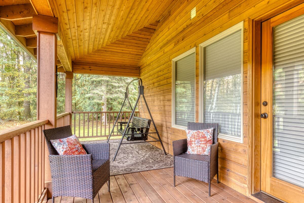 A covered wooden porch furnished with two dark wicker chairs featuring patterned cushions and a black metal swing bench. The porch overlooks a serene, dense green forest.