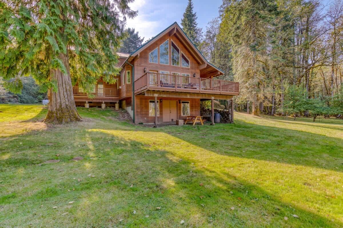 Side view of the wooden lodge featuring a large upper deck with a railing and expansive windows, overlooking a lush green lawn and surrounded by mature trees.