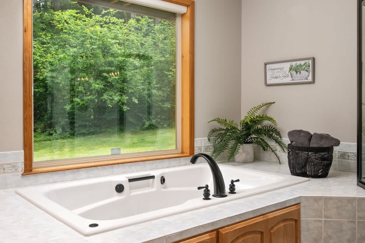 Close-up of a white built-in jetted bathtub with black fixtures, set into a light tiled counter. A large wooden-framed window offers a serene view of dense green trees.