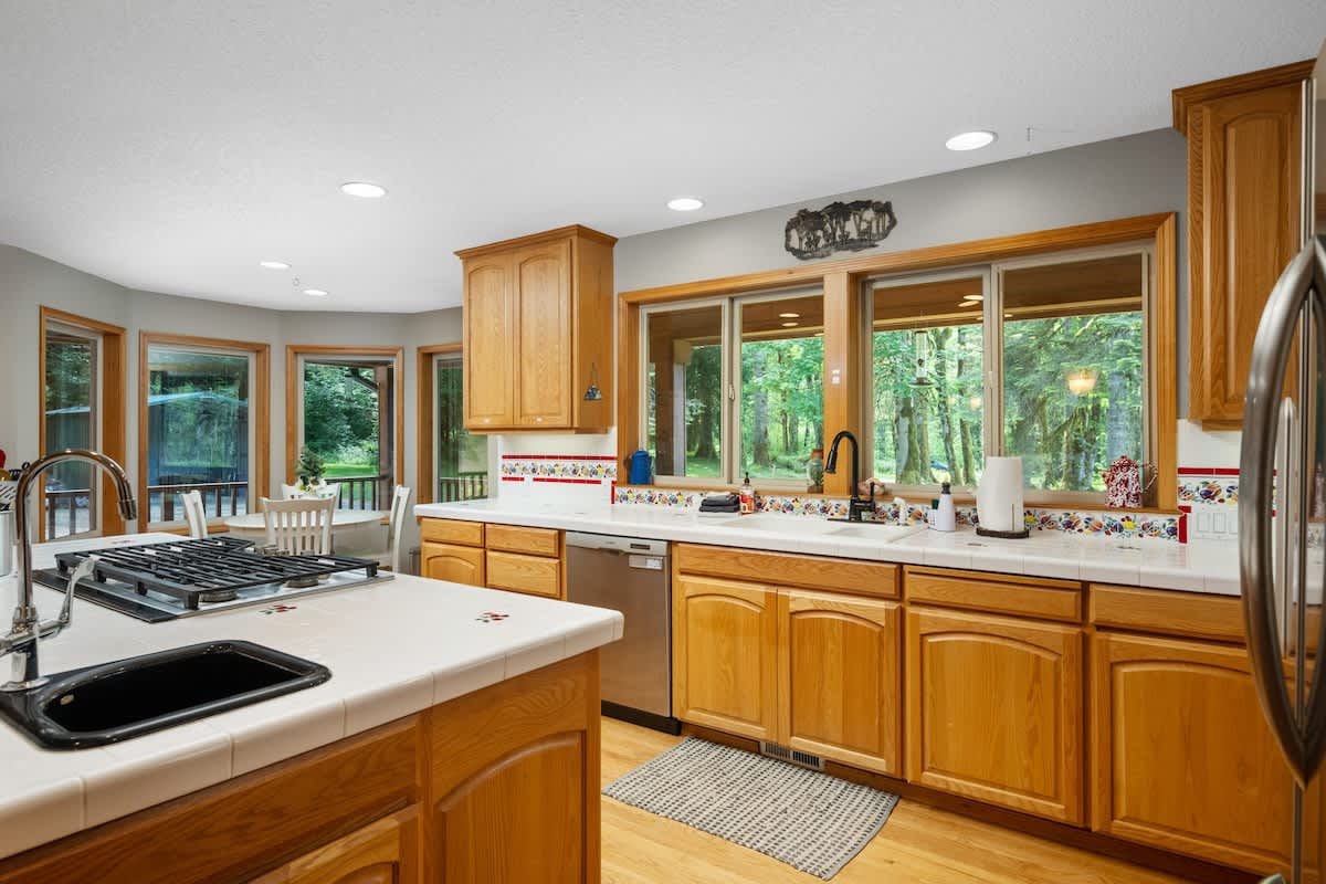Spacious kitchen with light wood cabinetry, white tiled countertops, and a large window overlooking a lush wooded area. Stainless steel appliances, including a dishwasher and refrigerator, are integrated into the design, with a gas cooktop on the island.