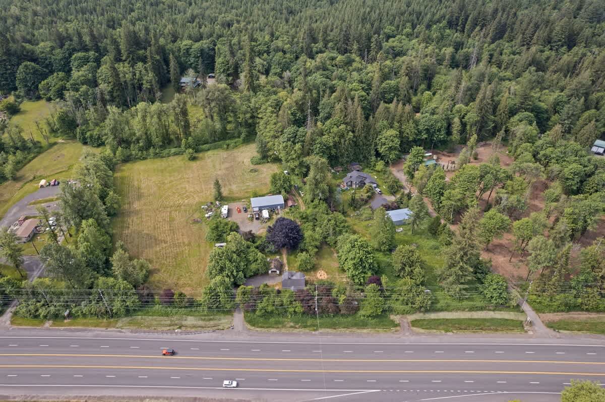 A wide aerial shot shows the property and surrounding residential areas amidst a vast forest, with a multi-lane highway running horizontally across the foreground, indicating its proximity to infrastructure.
