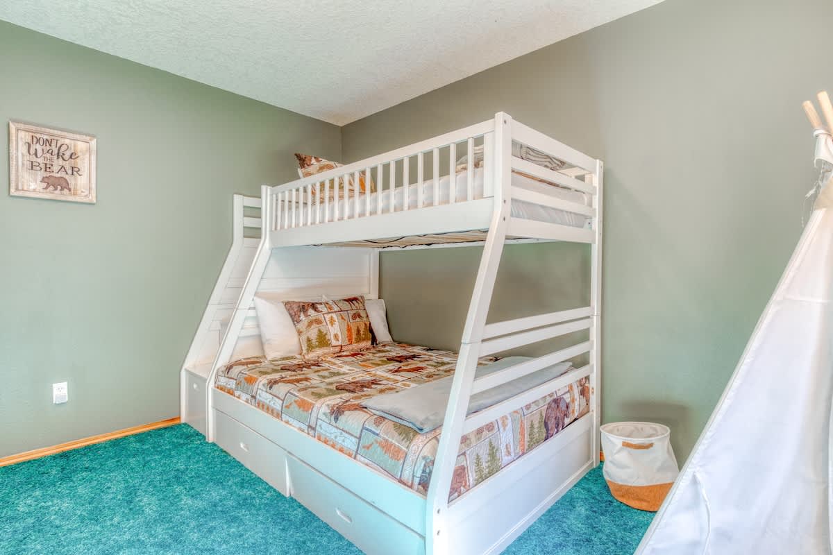 A cozy kids' bedroom with white wooden bunk beds, featuring a lower full bed and an upper twin, adorned with a bear-themed comforter, a white teepee, and a dark teal carpet.