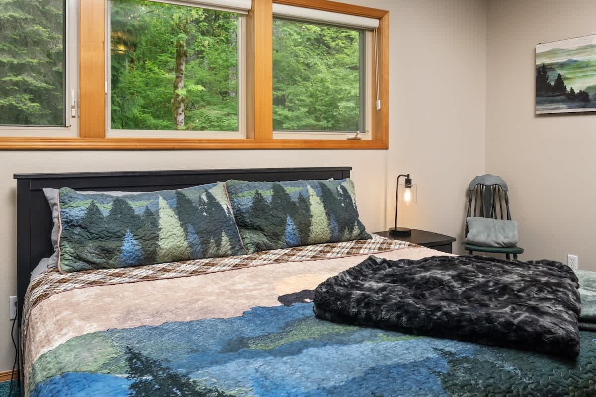 A close-up view of the bed in the bedroom, adorned with a blue and green patterned comforter and matching pillows. A black nightstand with a modern lamp is positioned beside the bed, with large windows in the background.